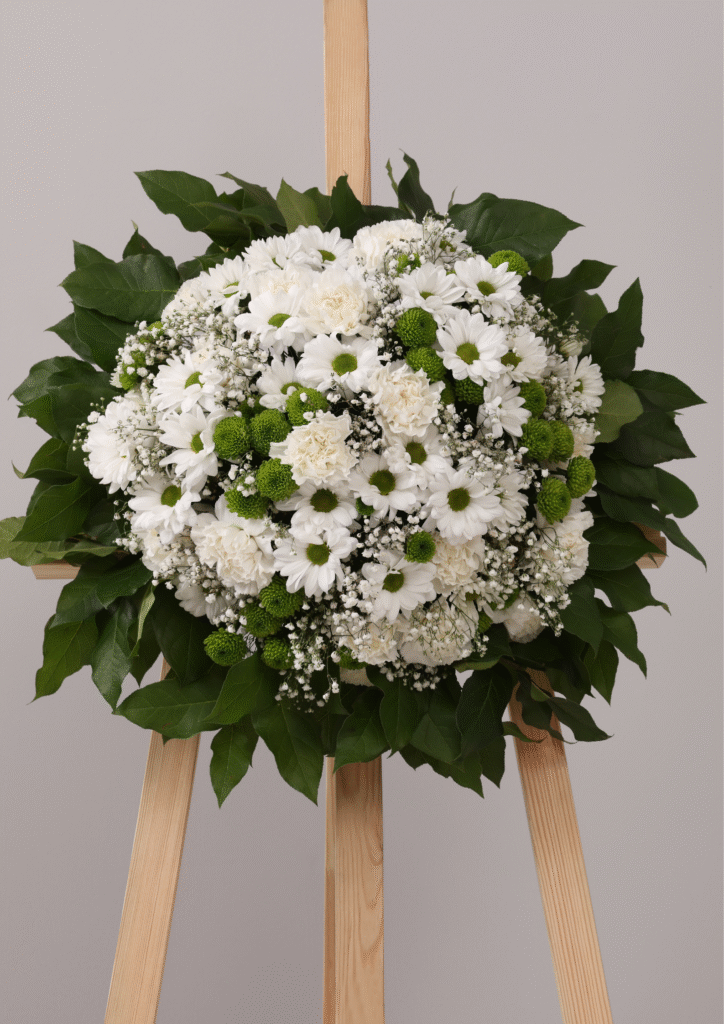 White funeral wreath with daisies, carnations, and greenery arranged on a wooden stand, symbolizing comfort and loving remembrance.