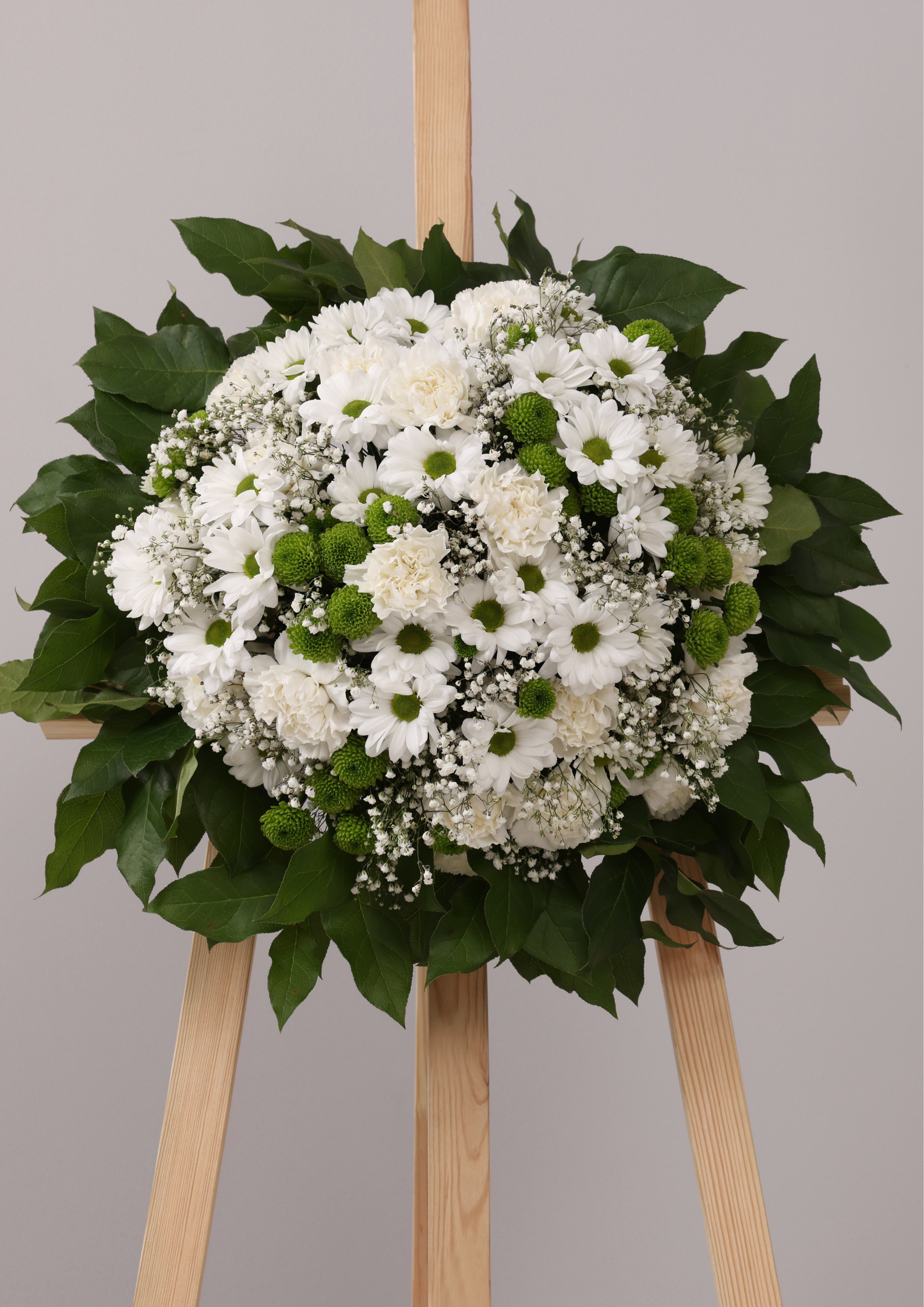 White funeral wreath with daisies, carnations, and greenery arranged on a wooden stand, symbolizing comfort and loving remembrance.